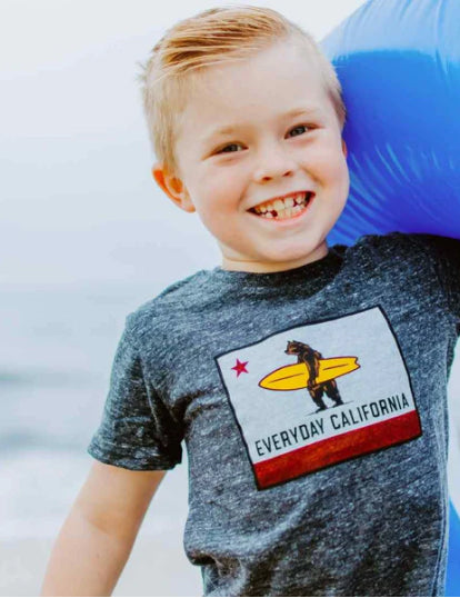 Photo of a young boy standing on the beach smiling directly at the camera. Everyday California Kids Apparel.